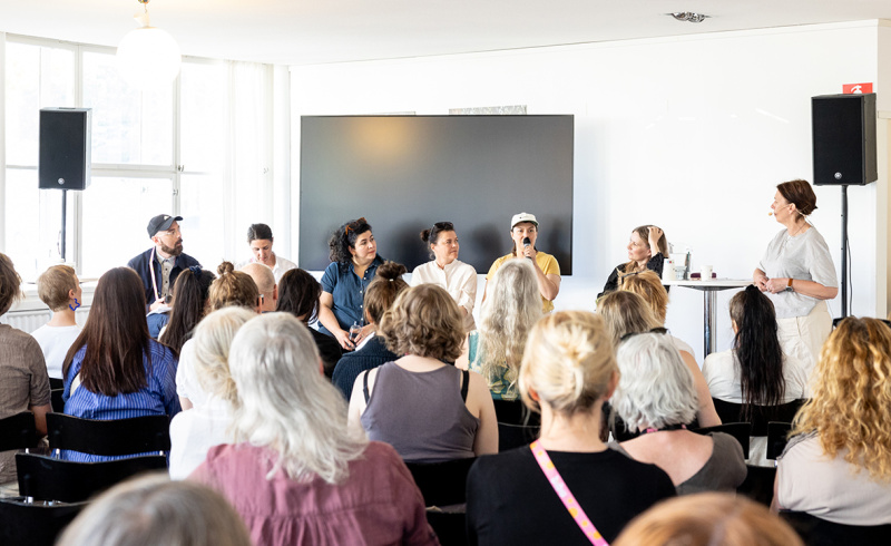Flera personer sitter bredvid varandra på en scen för att ge ett seminarier. Vi ser publiken bakifrån.